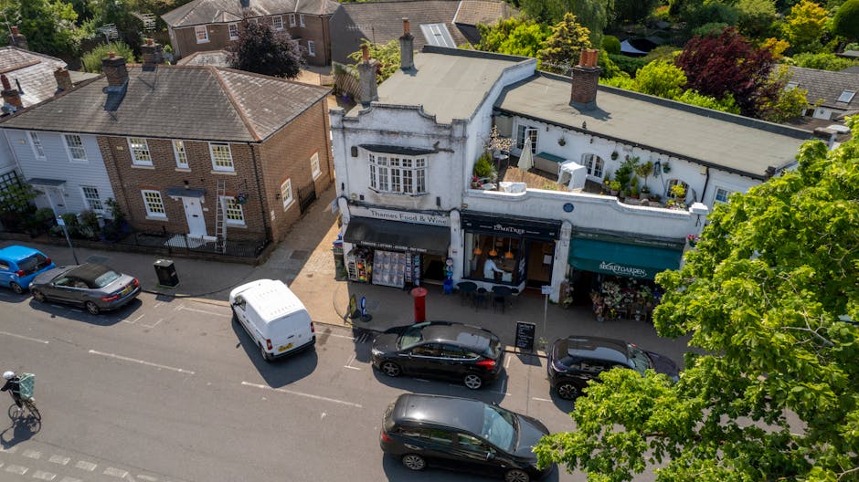 An aerial view of a street on Potters Bar High Street featuring a charming row of small shops with white and brick facades. In front of the shops, there are parked cars and a delivery van, indicating 