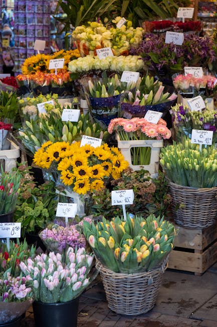 A colorful display of fresh flowers at a florist shop, featuring a variety of spring blooms such as yellow sunflowers, pink and white tulips, purple irises, and white roses. The flowers are arranged i