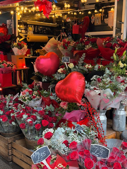 Inside a florist shop decorated for a romantic occasion, a variety of floral arrangements and bouquets are displayed on wooden crates and shelves. The arrangements predominantly feature red and white 