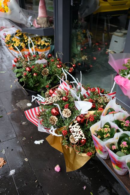 A colorful floral display outside a flower shop, featuring multiple festive bouquets arranged with a variety of flowers, including red and white carnations, pinecones, and red shiny baubles, decorated